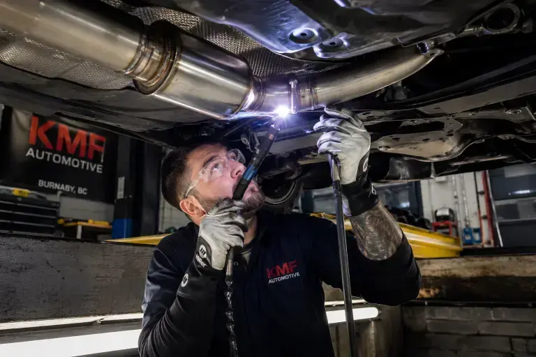An automotive technician welding a new exhaust pipe section beneath a car at KMF Automotive in Burnaby, BC.